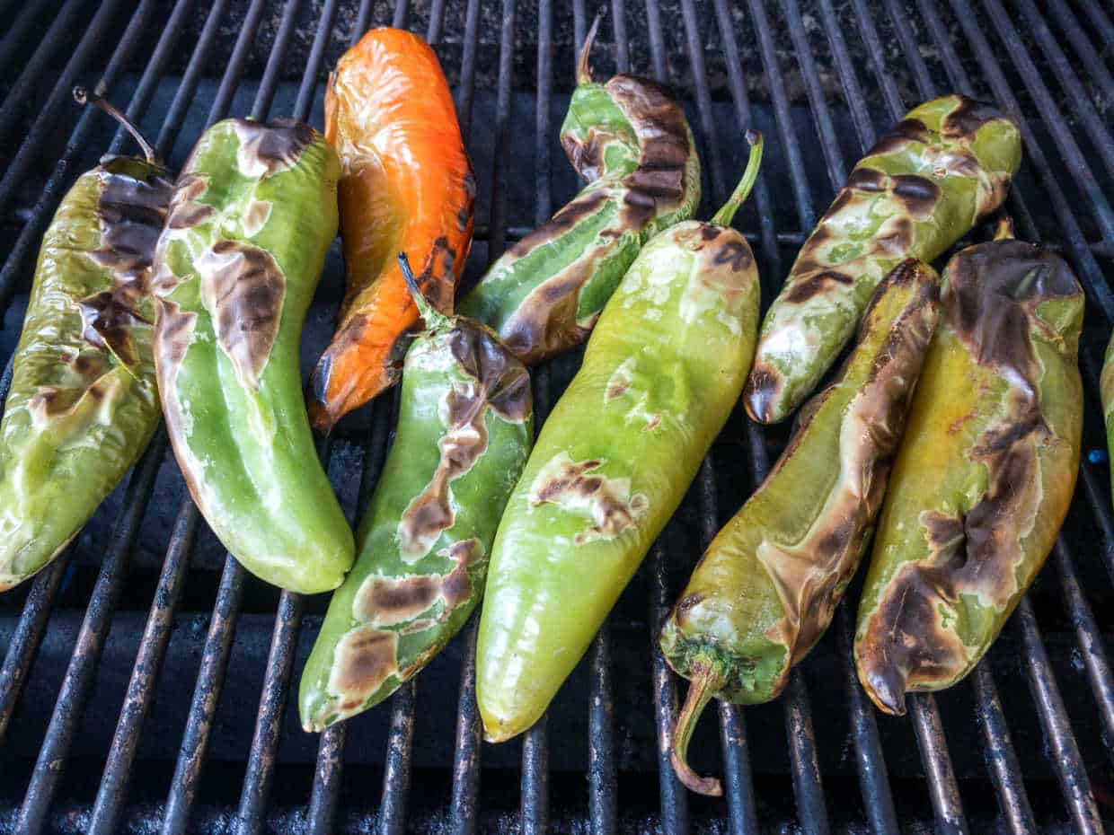 Several green and one orange Hatch chile peppers are being roasted on a grill, with charred and blistered skin visible on each pepper.