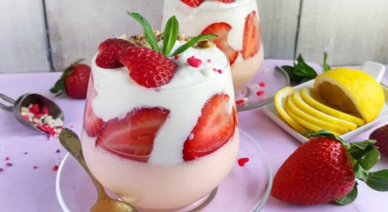 A glass of layered strawberry and cream dessert garnished with fresh strawberries, mint, and sprinkles, with lemon slices and whole strawberries beside it.