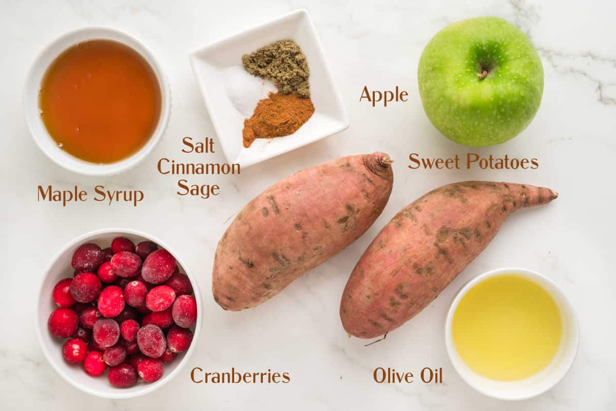 Ingredients on a white surface: maple syrup, salt, cinnamon, sage, a green apple, two sweet potatoes, cranberries, and olive oil.