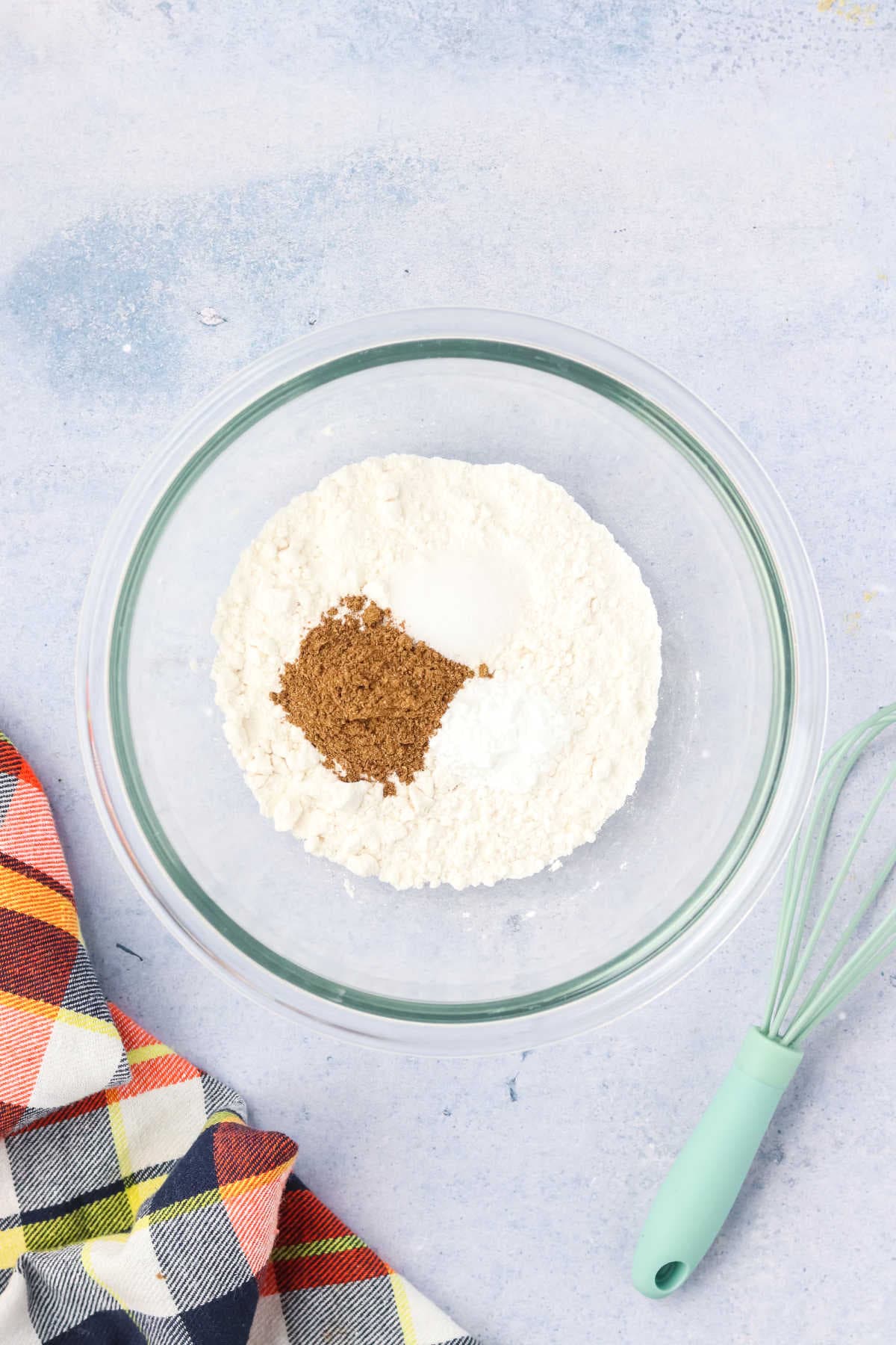 A glass bowl containing flour, brown sugar, and baking powder sits on a light surface next to a whisk and a plaid cloth.