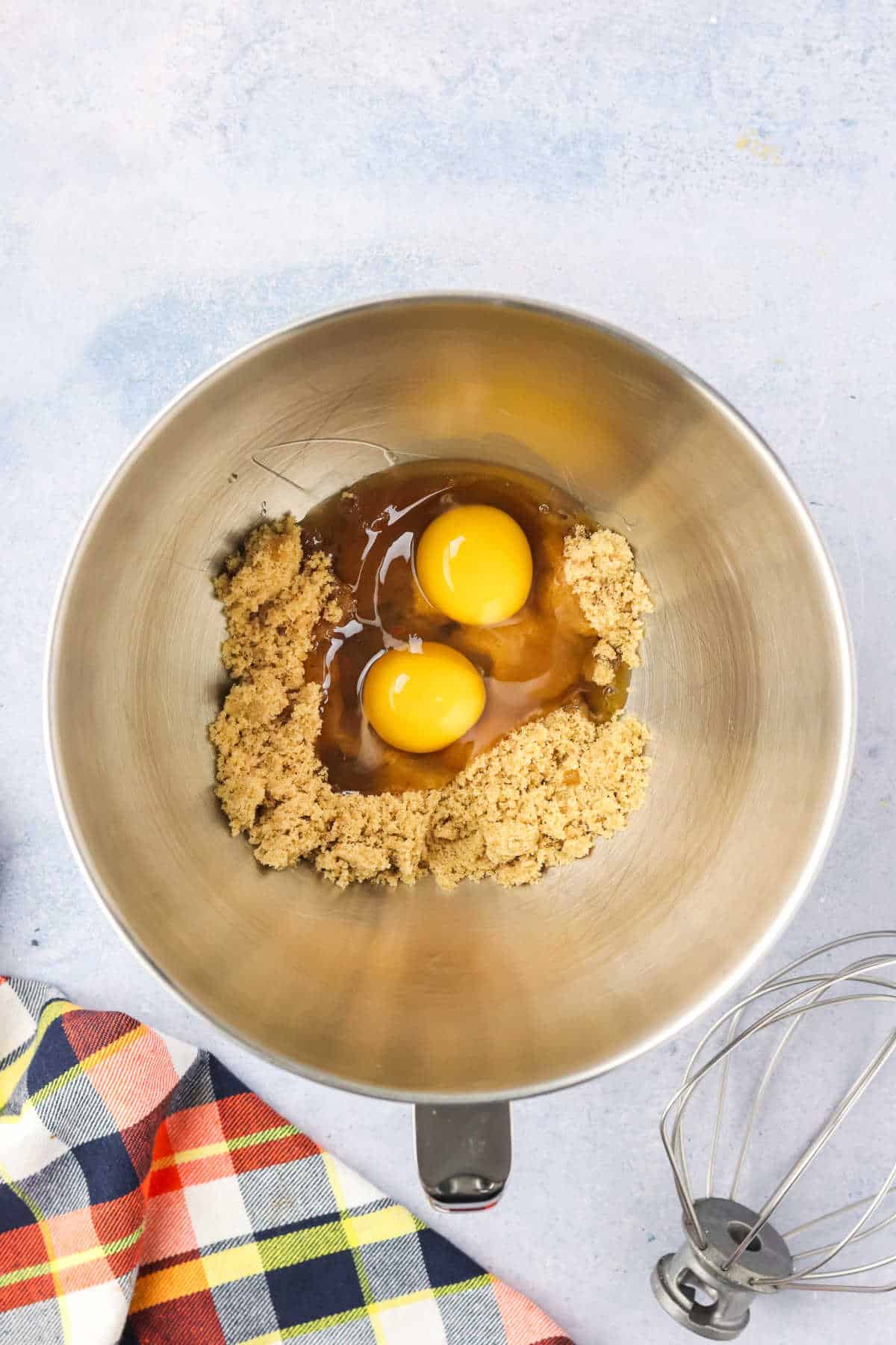A metal mixing bowl with brown sugar, two eggs, and vanilla extract, next to a wire whisk and a plaid cloth on a light surface.