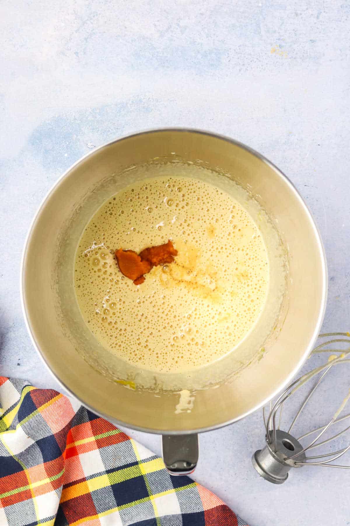 A mixing bowl containing a light yellow batter with pumpkin puree and vanilla on top, next to a whisk and a plaid cloth on a light-colored surface.