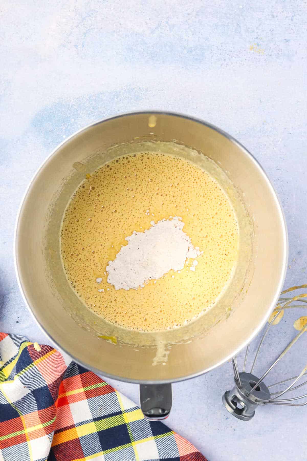 A metal mixing bowl containing a madelein cake batter with a mound of white flour on top, next to a whisk and a plaid cloth on a light surface.
