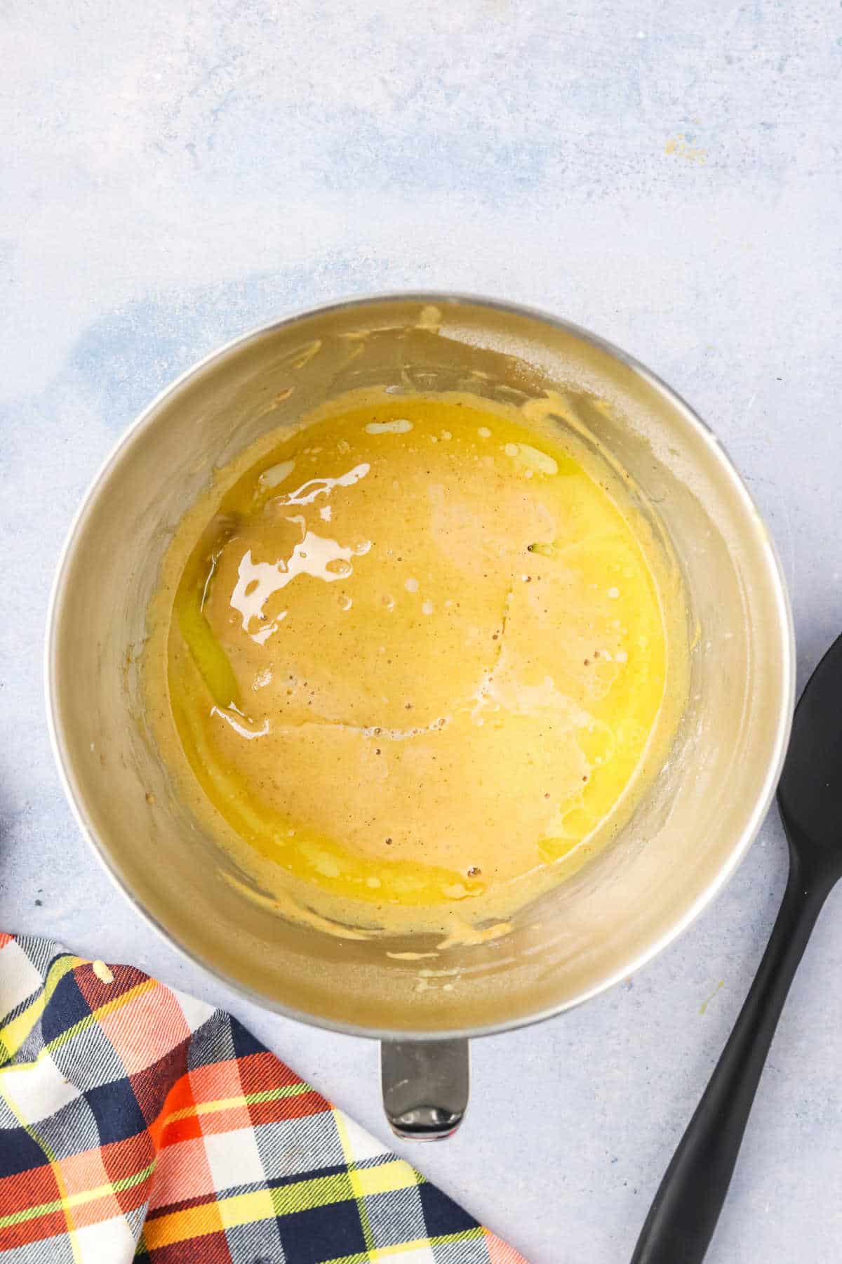 A stainless steel mixing bowl with a pumpkin madeleine batter inside sits melte butter next to a black spatula and a plaid cloth.
