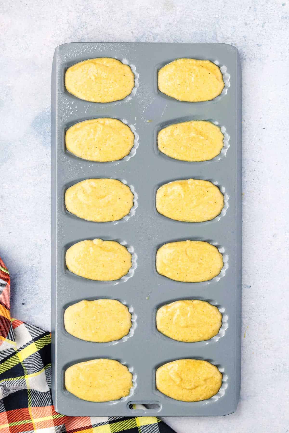 A gray madeleine baking pan filled with pumpkin batter, ready to be baked, sits on a light countertop next to a plaid cloth.