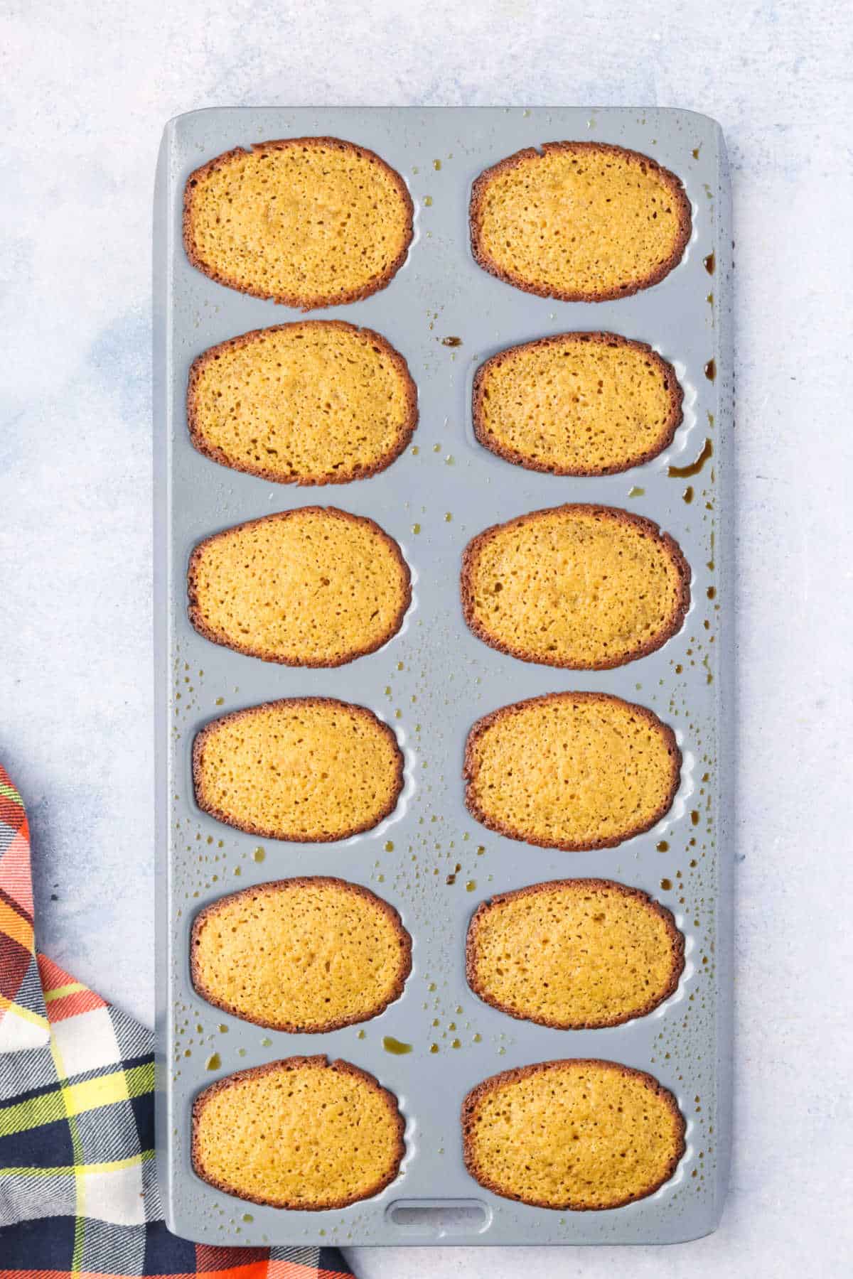 A madeleine pan with twelve freshly baked pumpkin madeleines, still in the mold, on a light surface next to a checked cloth.