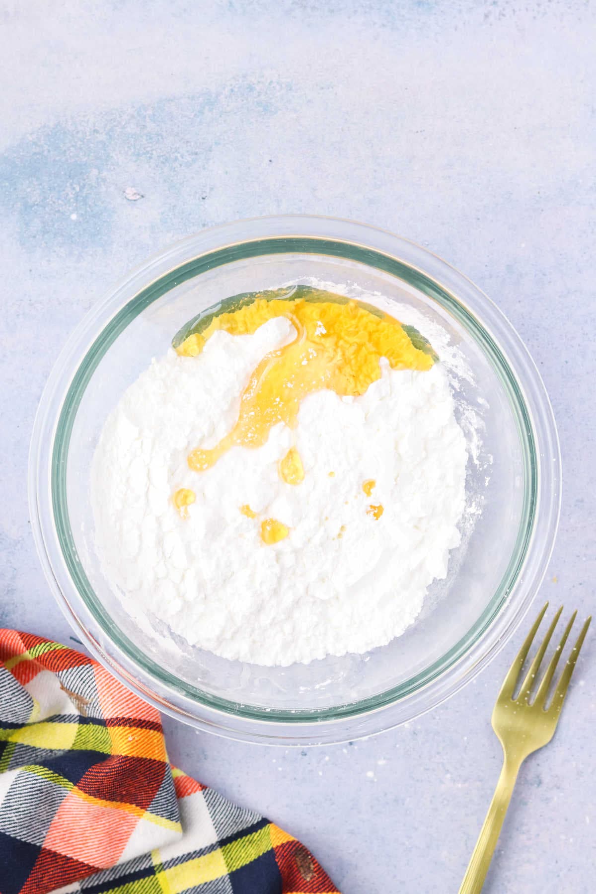 A glass bowl containing powdered sugar and melted butter, with a gold fork and a plaid cloth nearby on a light surface.