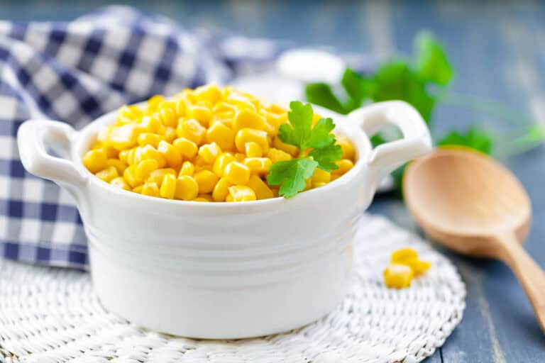 A white ceramic bowl filled with cooked corn kernels, garnished with parsley, sits on a woven mat beside a wooden spoon and a checkered cloth.