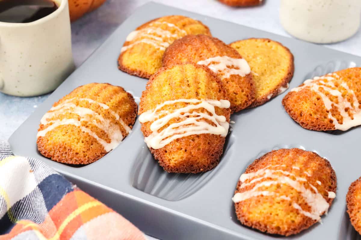A baking pan holds several pumkin madeleines, some drizzled with white icing, next to a mug and a plaid cloth.