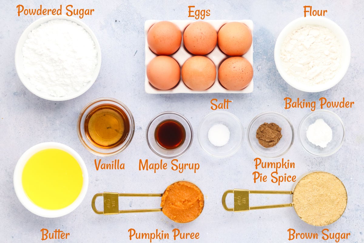 Ingredients for baking are arranged on a countertop, including eggs, flour, powdered sugar, butter, pumpkin puree, brown sugar, vanilla, maple syrup, salt, baking powder, and pumpkin pie spice.