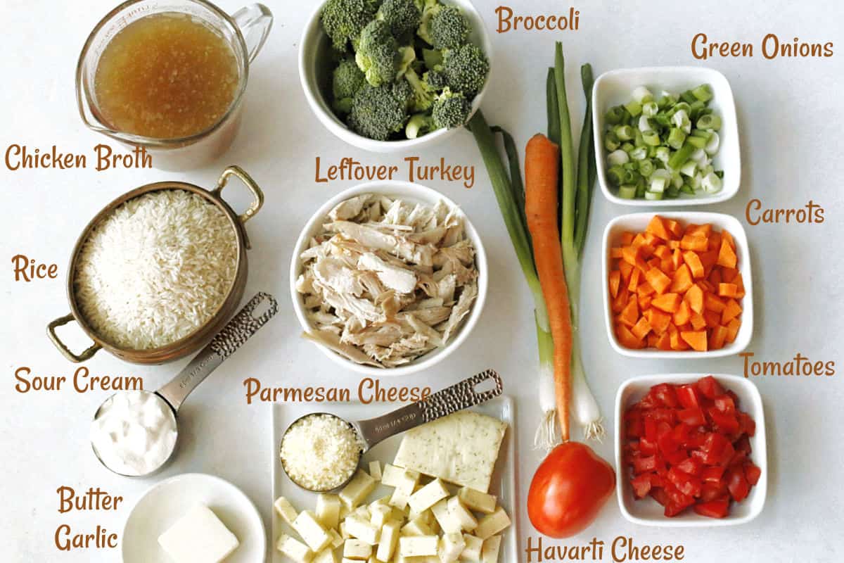 Labeled ingredients for a leftover turkey and rice casserole are arranged on a white surface, including chicken broth, rice, broccoli, green onions, carrots, tomatoes, turkey, parmesan, havarti cheese, sour cream, butter, and garlic.