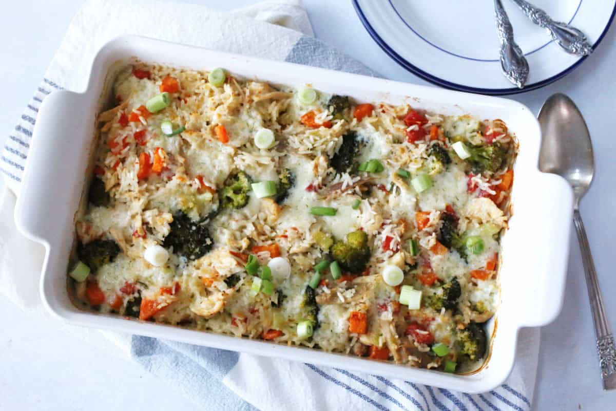 A white baking dish filled with a baked turkey casserole containing rice, broccoli, diced red peppers, melted cheese, and chopped green onions sits on a striped towel next to plates and utensils.