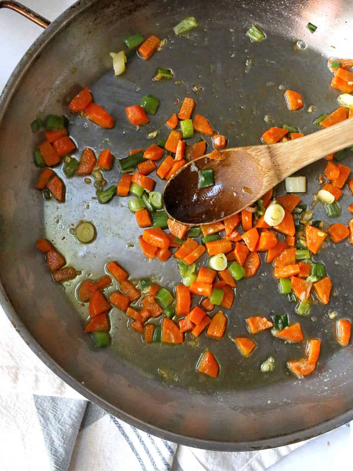 Chopped carrots, green onions, a saut&eacute;ing in a large pan with a wooden spoon on top.