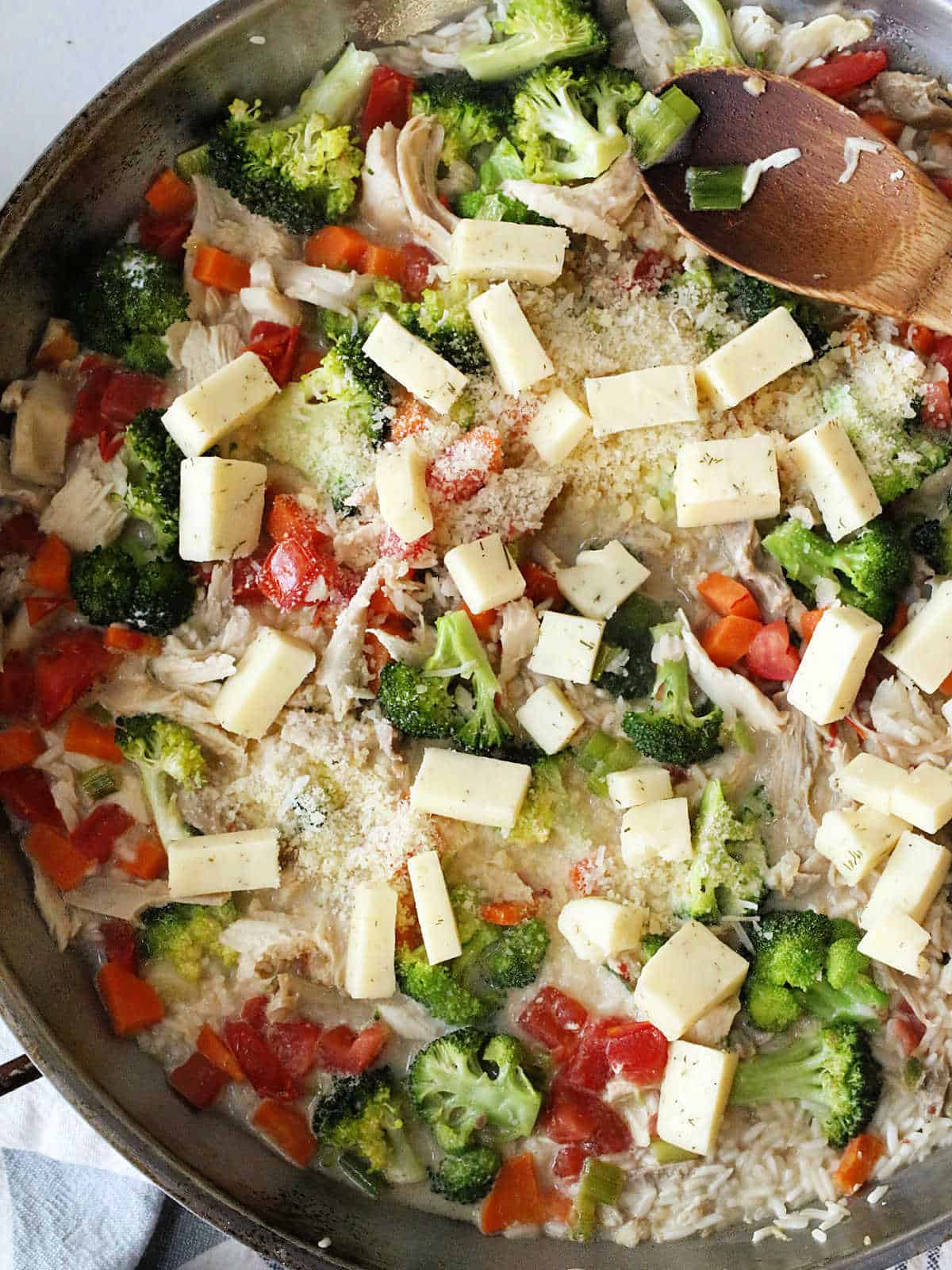A skillet filled with cooked rice, broccoli, shredded turkey, diced tomatoes, carrots, and cubed cheese, being stirred with a wooden spoon.