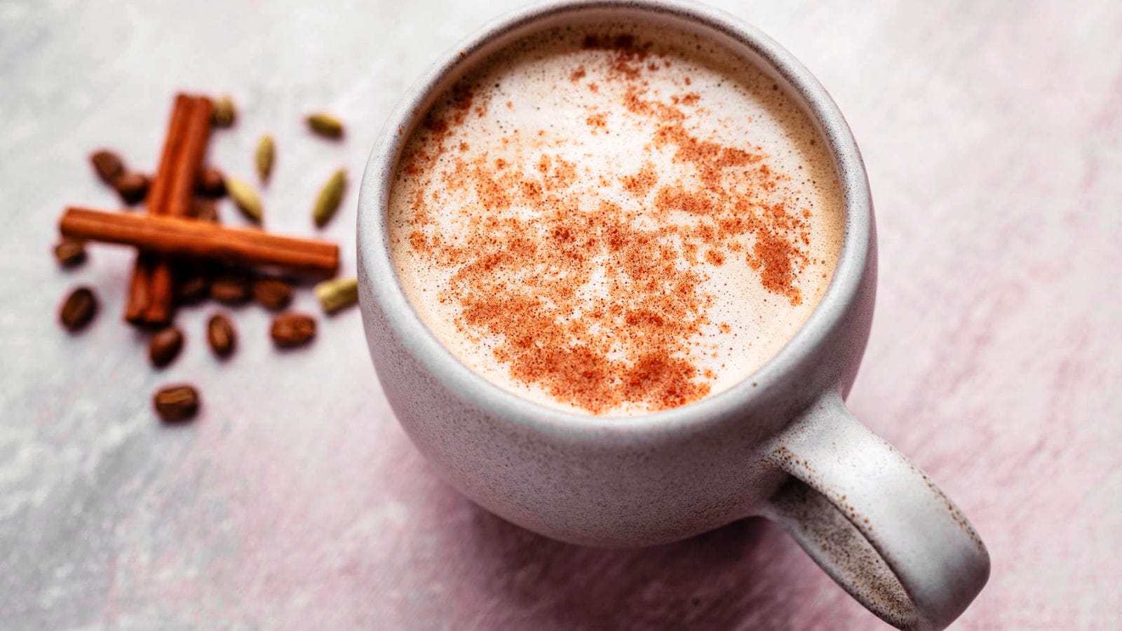 A ceramic mug filled with a frothy beverage topped with cinnamon, next to cinnamon sticks, cardamom pods, and coffee beans on a light pink surface.