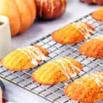 Golden brown madeleines with white icing are cooling on a wire rack, with pumpkins and a cup of coffee visible in the background.