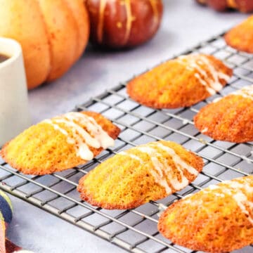 Golden brown madeleines with white icing are cooling on a wire rack, with pumpkins and a cup of coffee visible in the background.