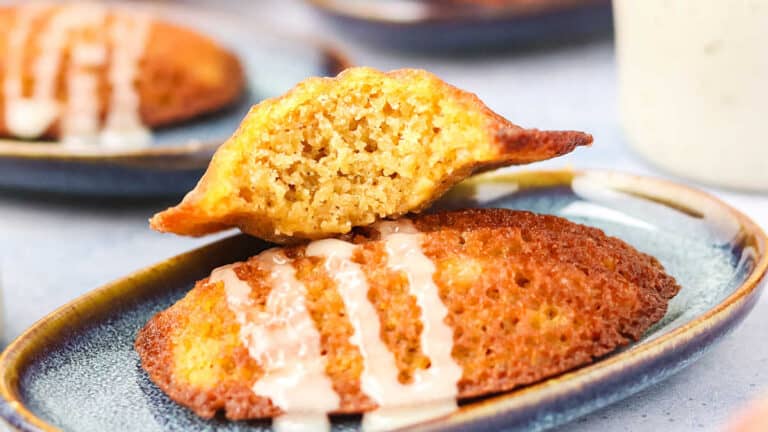 Two golden brown madeleines on a ceramic plate, one whole and one halved, with white icing drizzled on top.