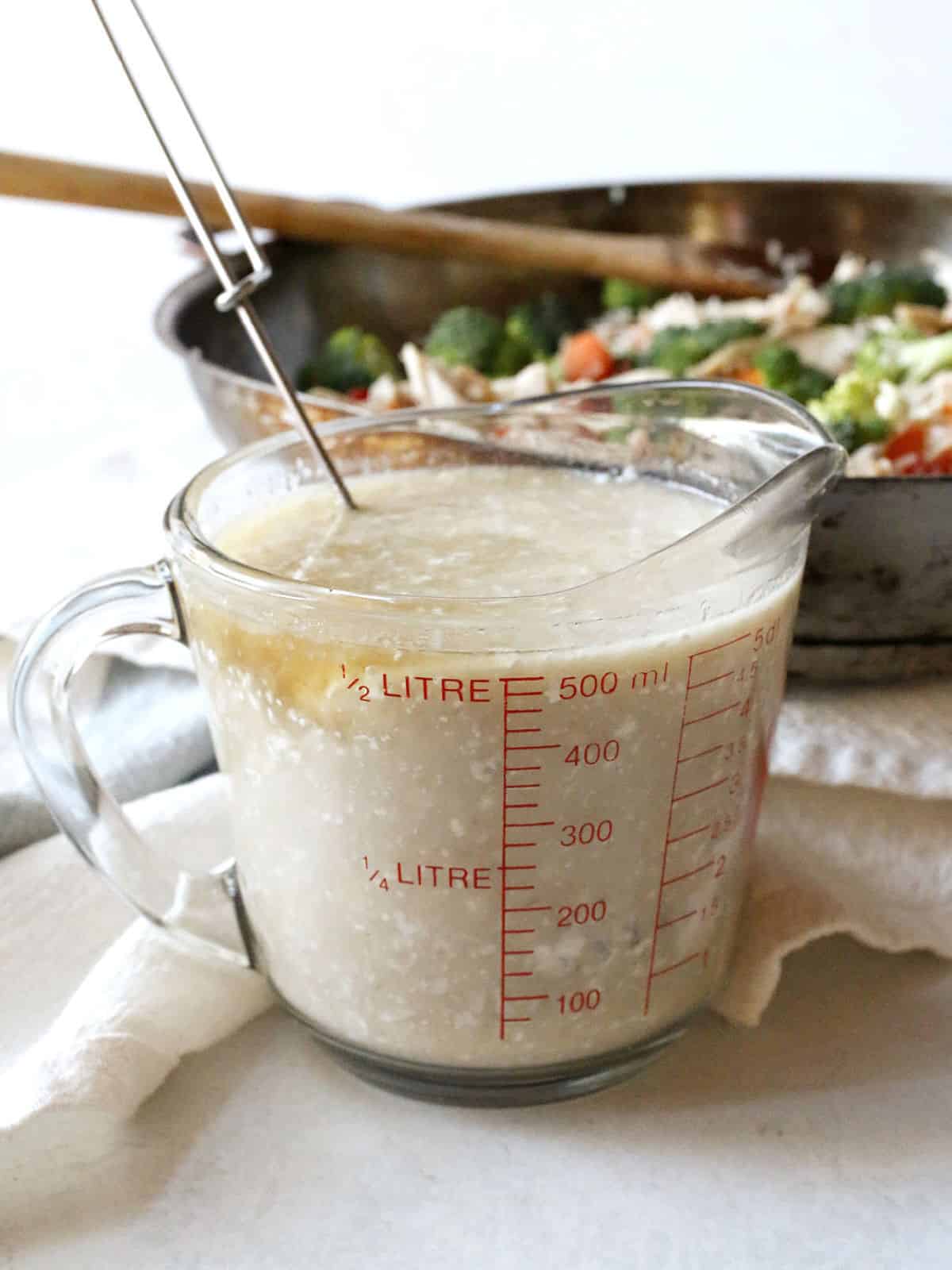 A glass measuring cup filled with a chicken broth and sour cream sits on a white cloth, with a metal utensil inside and a skillet of vegetables in the background.