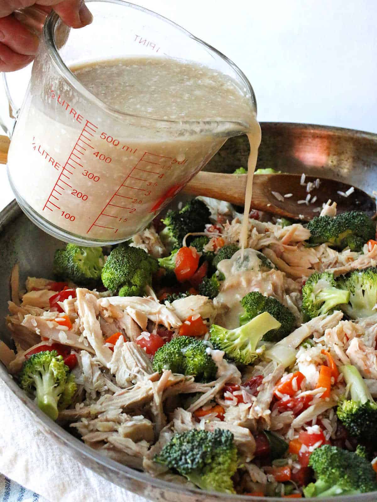 A hand pours creamy sauce from a measuring cup over a skillet filled with shredded turkey, broccoli, red bell peppers, and rice.
