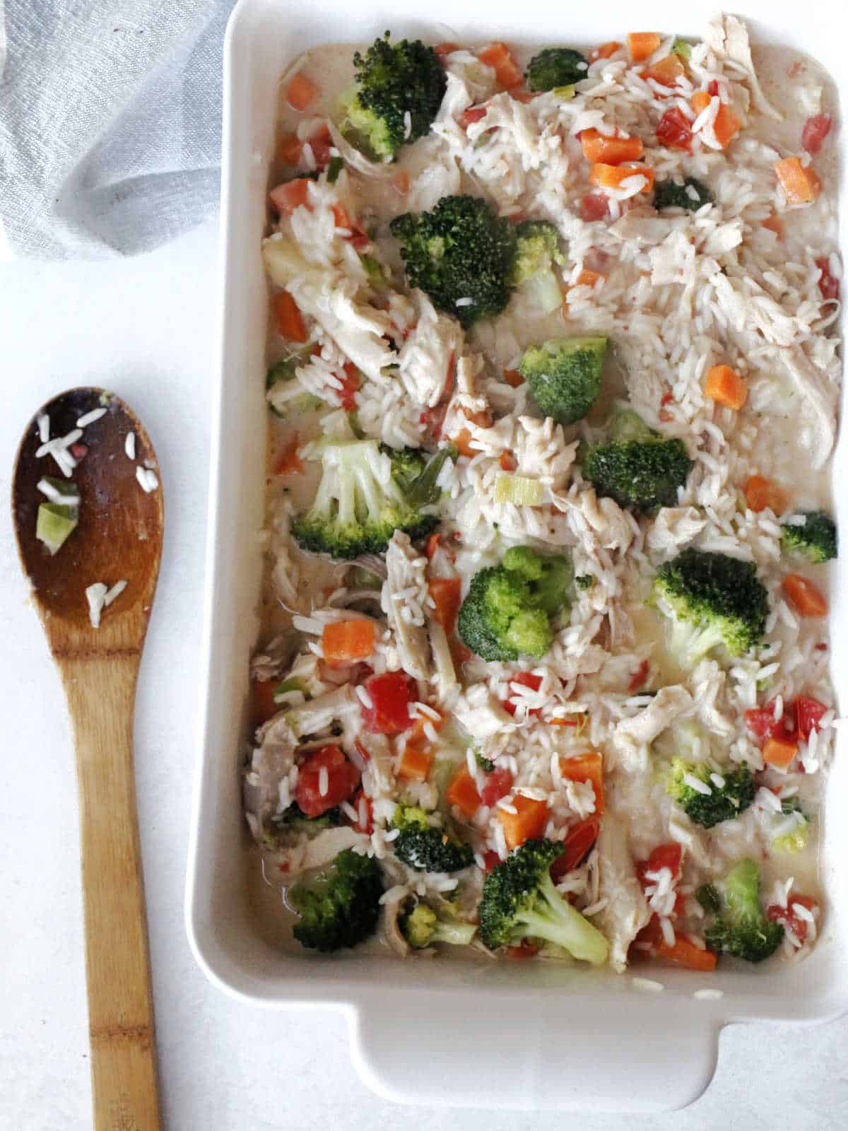 A white baking dish filled with a mixture of rice, shredded turkey, broccoli, diced carrots, and red bell peppers. A wooden spoon with rice residue lies next to the dish.