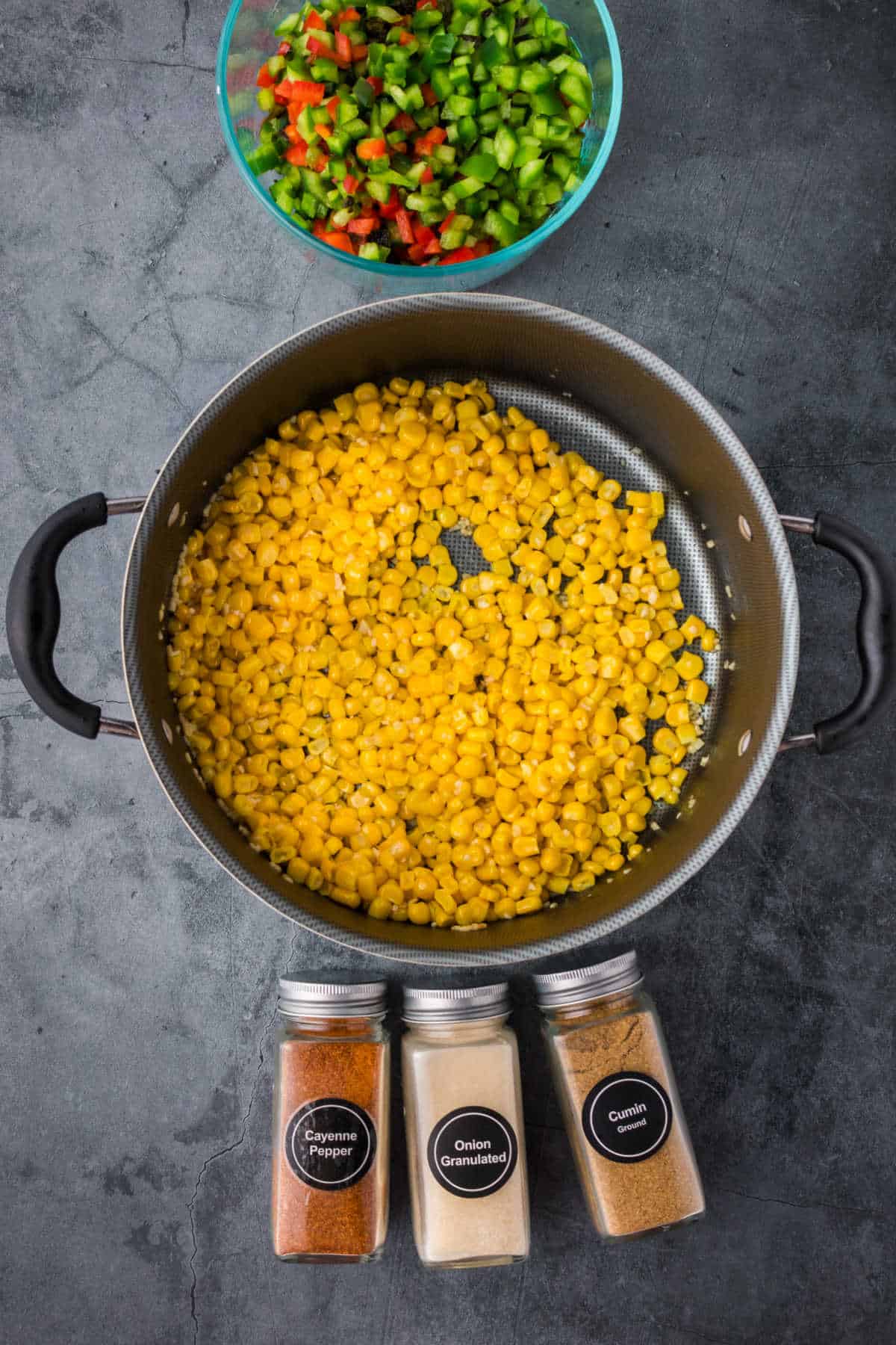 Pan with cooked corn, three spice containers labeled cayenne pepper, onion granulated, and cumin, and a bowl of chopped green and red bell peppers on a dark surface.