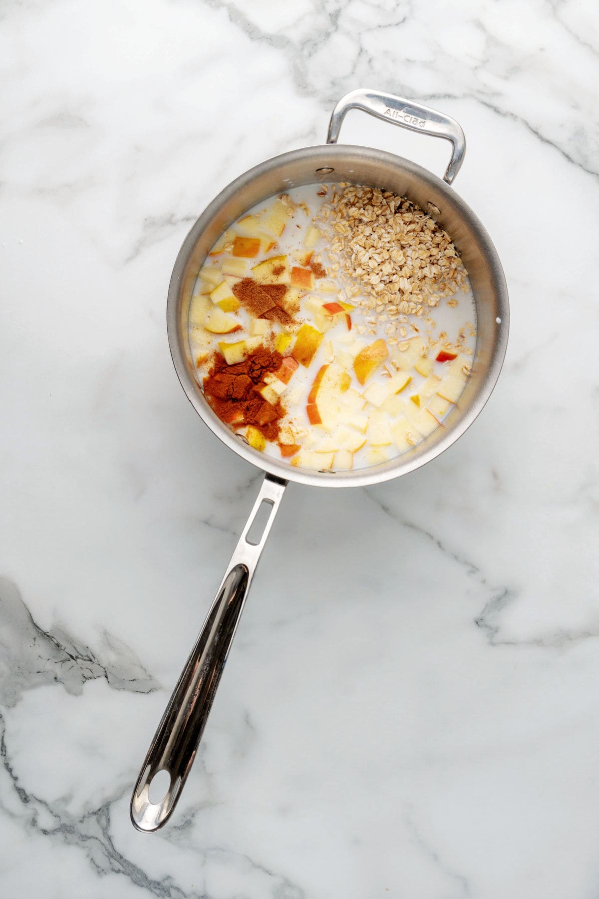 A saucepan with milk, chopped apples, oats, and spices on a marble countertop, ingredients not yet mixed for old-fashioned oatmeal.
