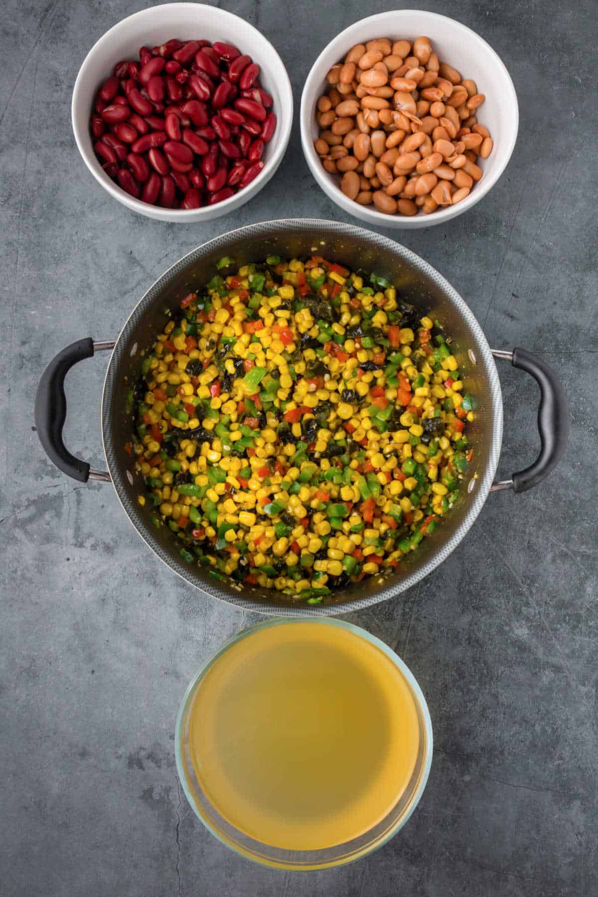 A pot of mixed vegetables sits on a gray surface, with separate bowls of kidney beans, pinto beans, and vegetable broth surrounding it.