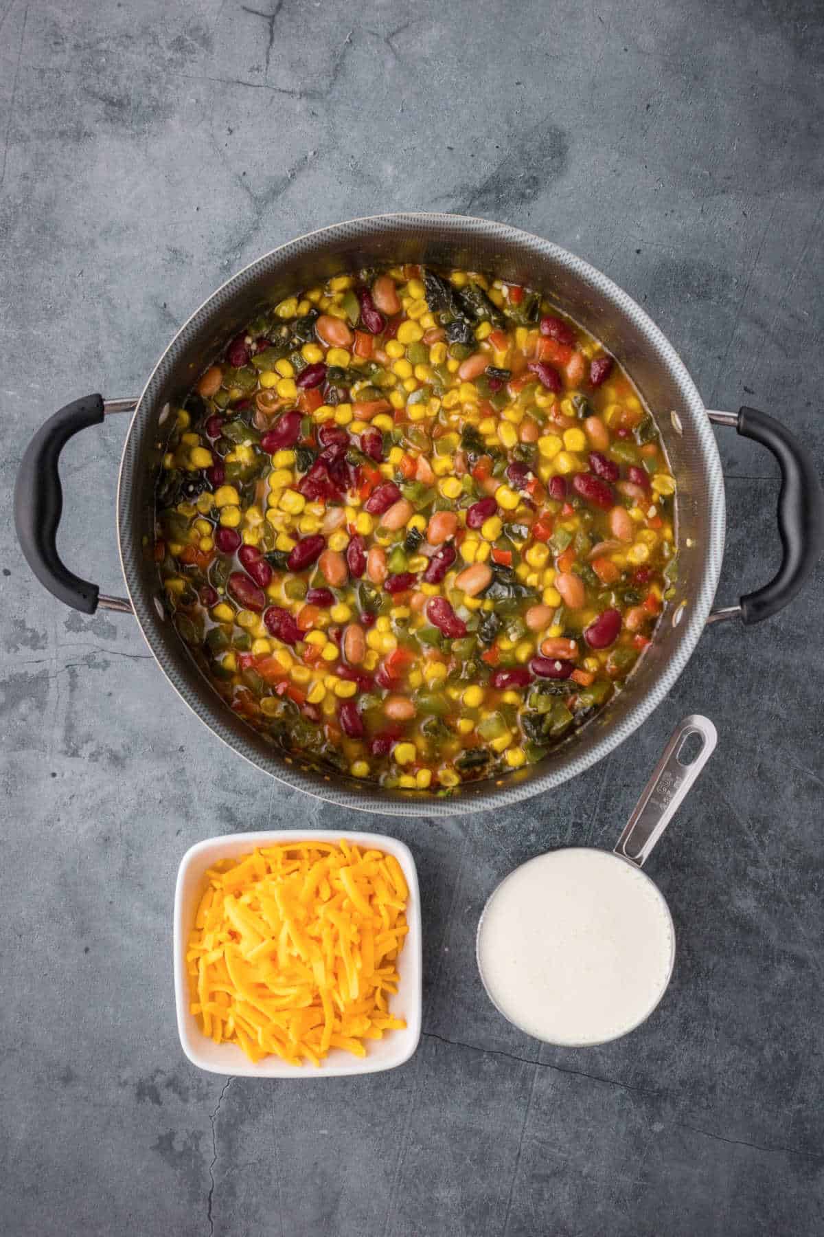 A pot of mixed vegetable and bean soup sits on a gray surface next to a bowl of shredded cheddar cheese and a measuring cup of cream.