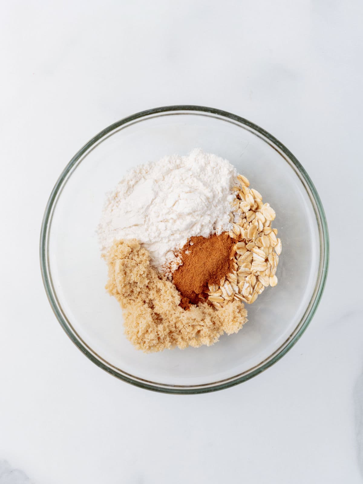 A glass bowl containing brown sugar, flour, rolled oats, and ground cinnamon on a white surface.