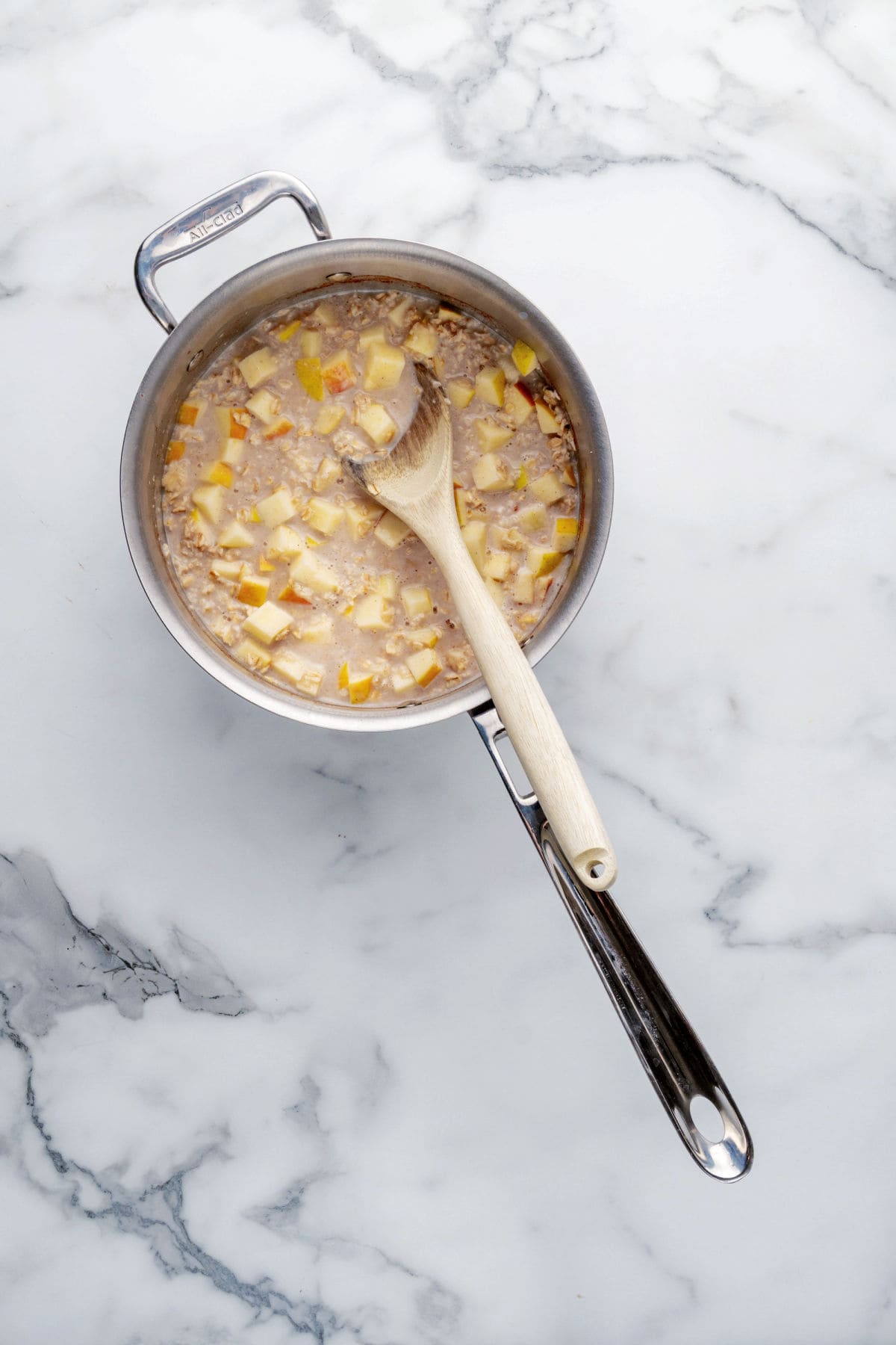 A saucepan with chopped apples and oats being cooked and stirred with a wooden spoon, placed on a marble countertop.