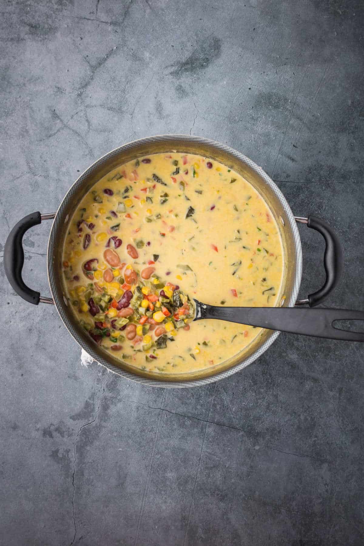 A pot of creamy Mexican bean soup with corn, herbs, and vegetables, shown from above on a gray countertop with a black ladle resting inside.