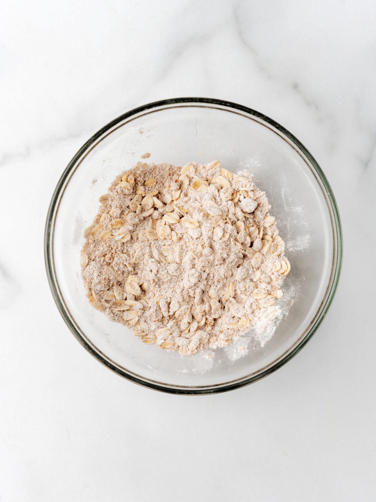 A glass bowl filled with a mixture of oats, flour, and brown sugar sits on a white marble surface.