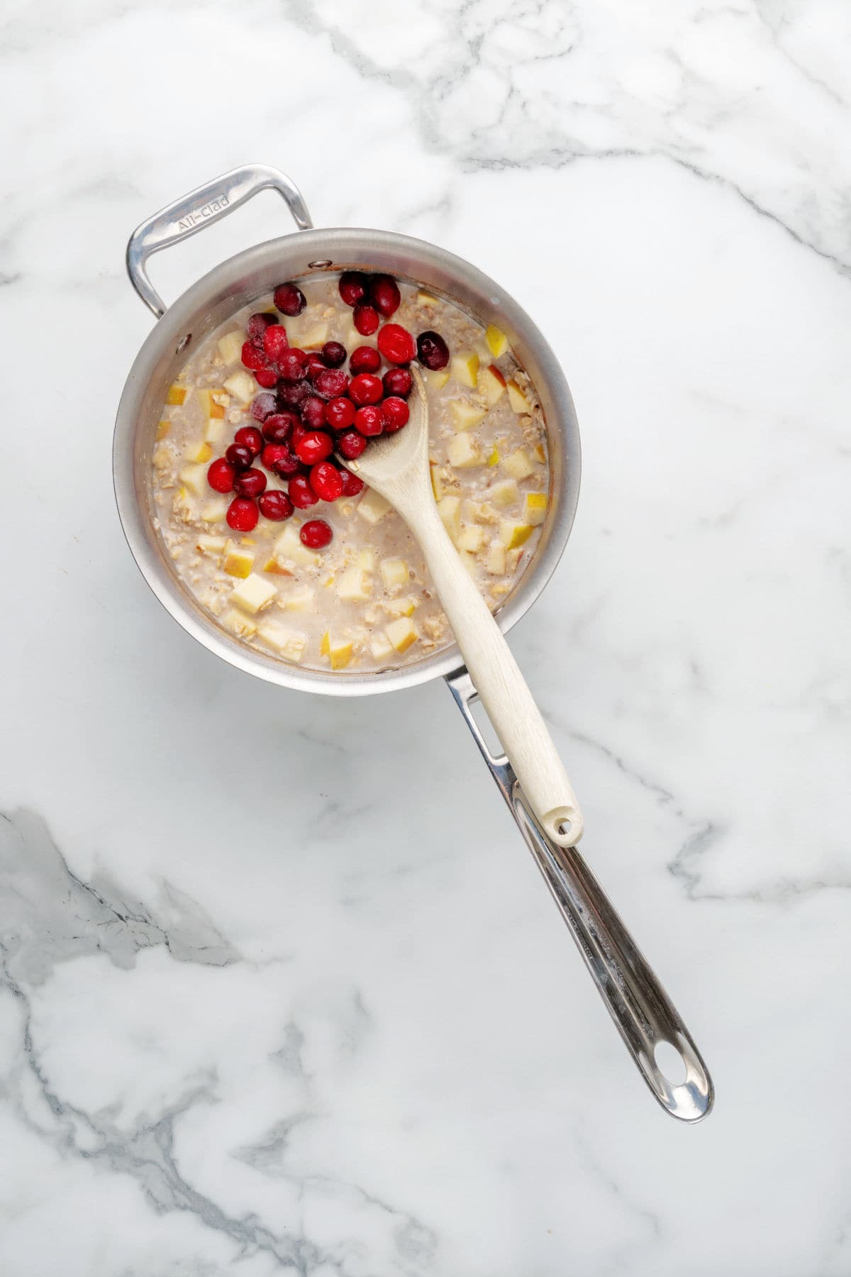 A pot of oatmeal with diced fruit and fresh cranberries being stirred with a wooden spoon on a marble countertop.
