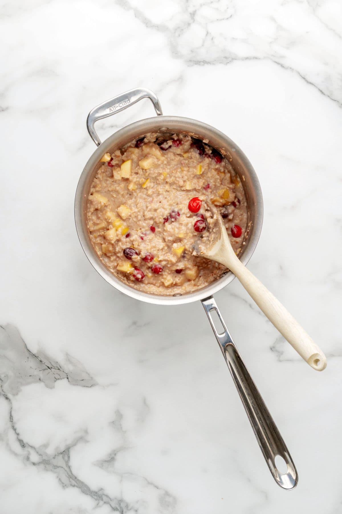 A saucepan filled with oatmeal mixed with apples and cranberries sits on a marble surface; a wooden spoon rests inside the pot.