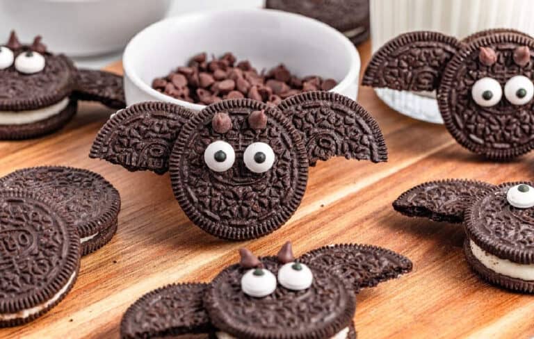 Chocolate sandwich cookies decorated with candy eyes and broken cookie wings to resemble bats, displayed on a wooden surface with a bowl of chocolate chips in the background.