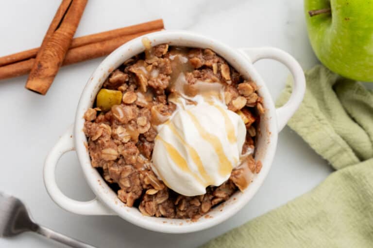 A small white dish of apple crisp topped with a scoop of vanilla ice cream and caramel sauce, next to a green apple, cinnamon sticks, and a green cloth.