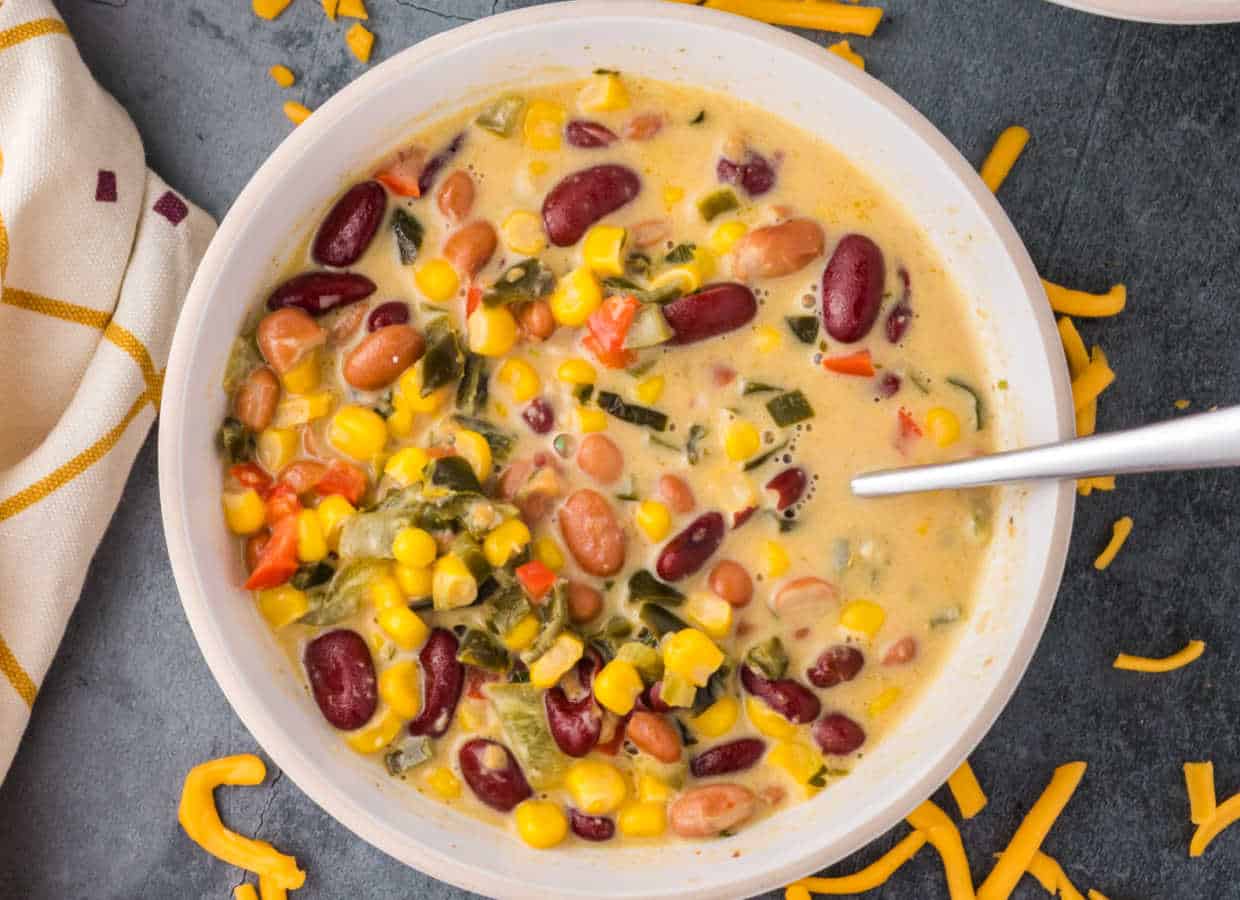 A bowl of creamy soup with kidney beans, pinto beans, corn, diced vegetables, and a spoon, with shredded cheese scattered nearby.