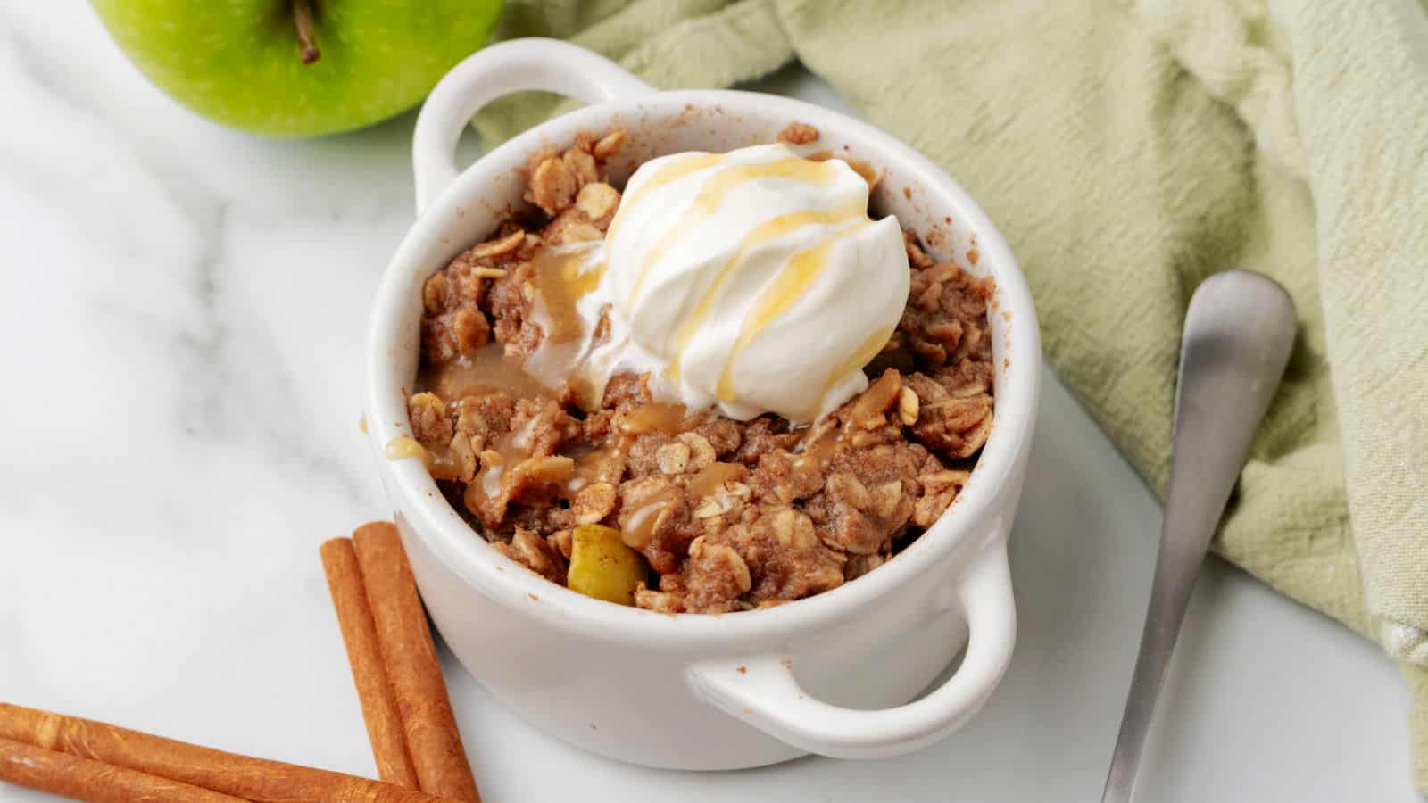 A white ramekin filled with apple crisp, topped with whipped cream and caramel drizzle, sits on a marble surface with a spoon, cinnamon sticks, an apple, and a green cloth nearby.