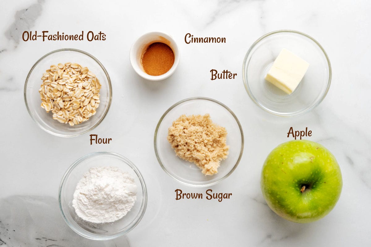 A green apple, flour, old-fashioned oats, brown sugar, butter, and cinnamon are arranged in bowls on a white surface for apple crisp in a mug.