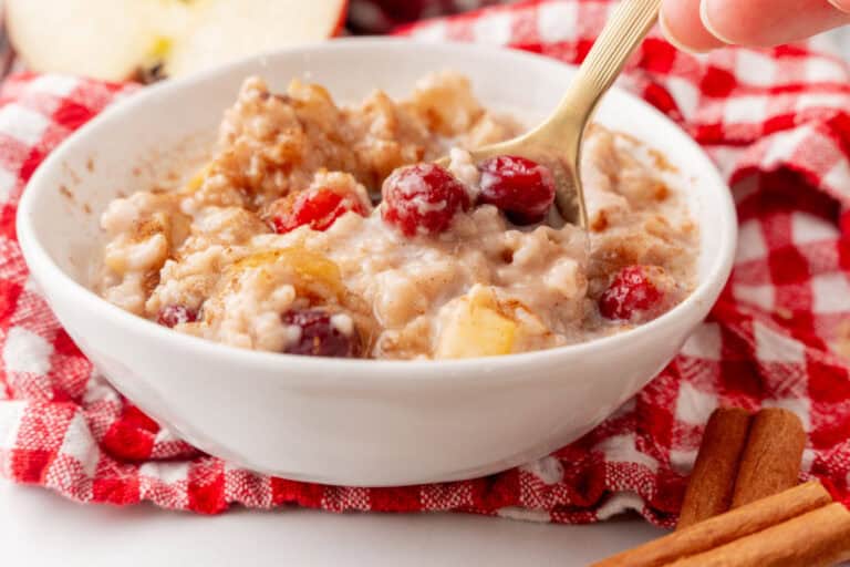 A bowl of oatmeal with cranberries and apples on a red checkered cloth, with a spoon and cinnamon sticks nearby.