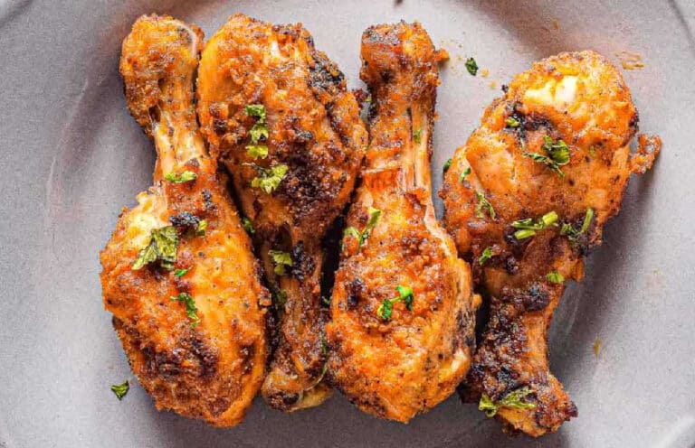 Four seasoned and cooked chicken drumsticks garnished with chopped herbs are arranged on a grey plate.