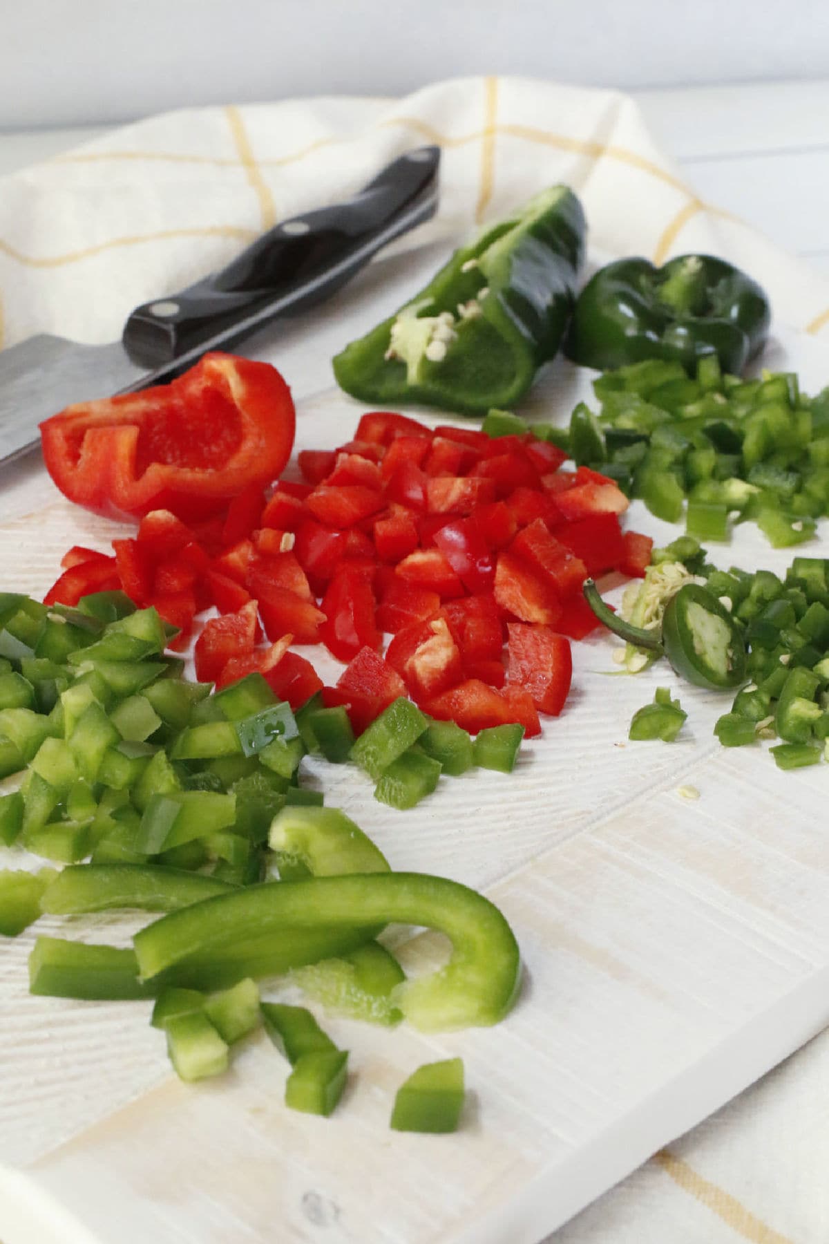 Chopped red and green bell peppers on a white cutting board with a knife and uncut peppers in the background.