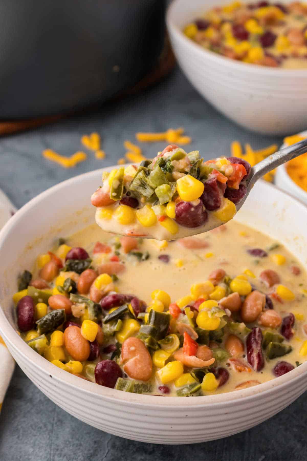 A bowl of creamy soup with beans, corn, diced vegetables, and shredded cheese, with a spoonful being lifted from the bowl.