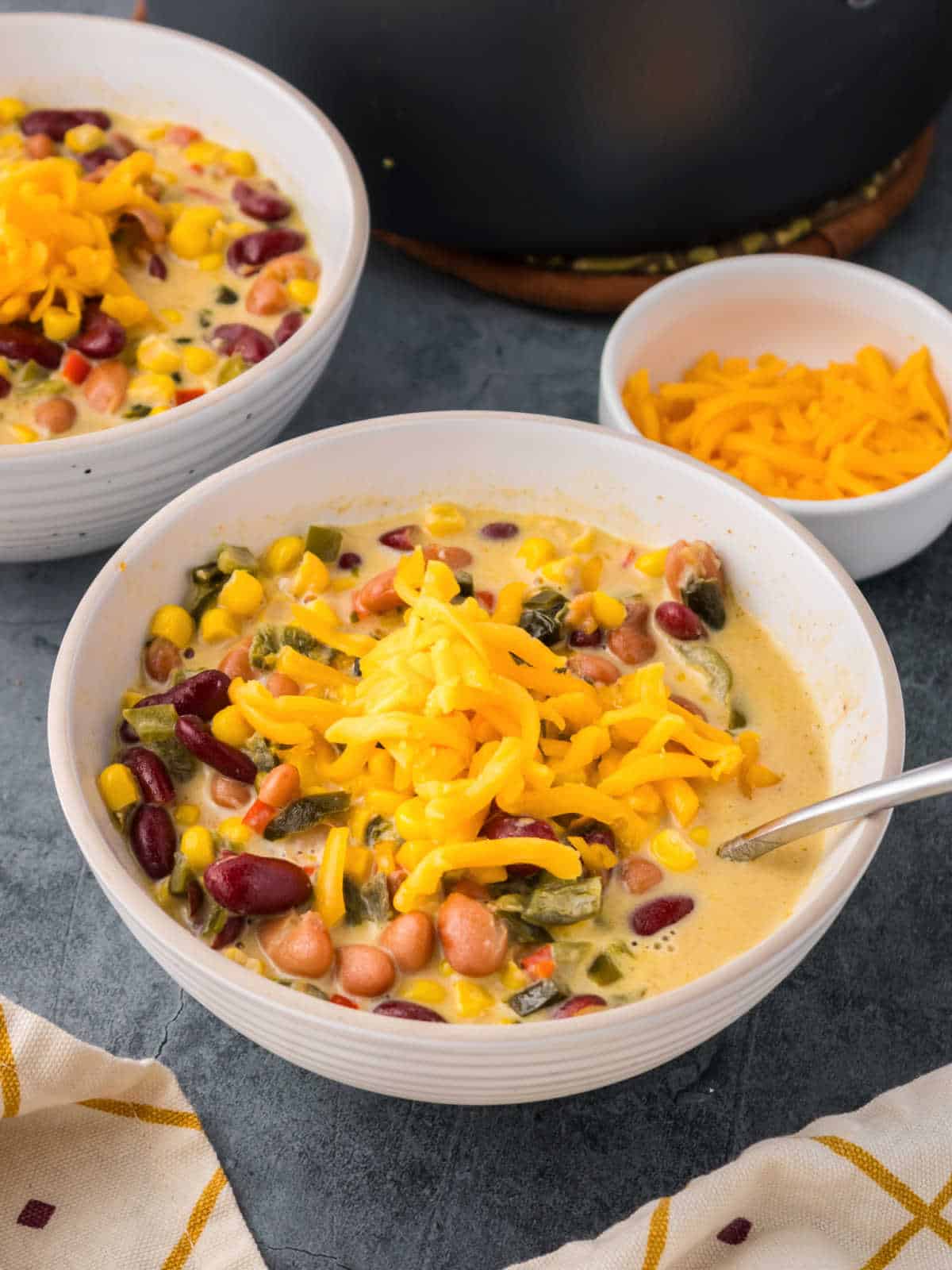 A bowl of creamy soup with beans, corn, and chopped vegetables, topped with shredded cheddar cheese, with a spoon and extra cheese in a small bowl nearby.