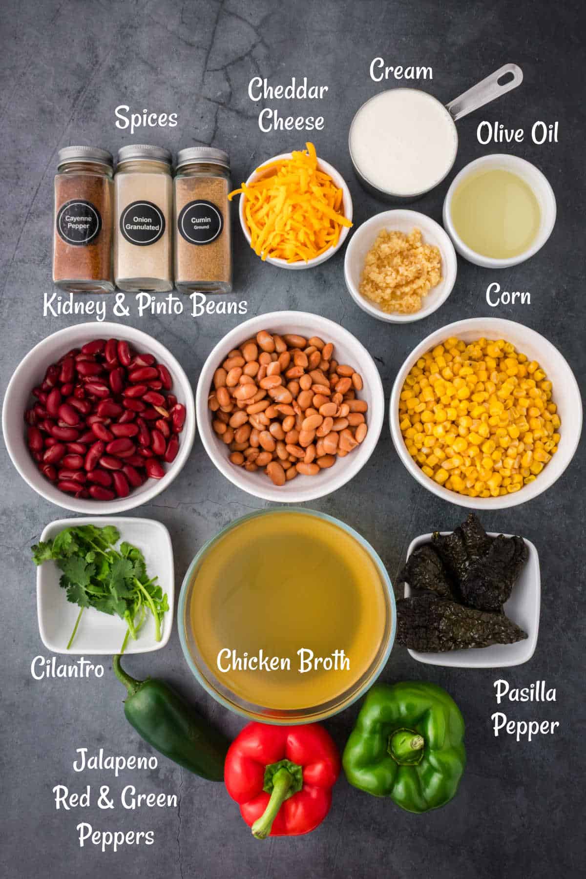 Assorted ingredients for a soup recipe, including spices, cheddar cheese, cream, olive oil, garlic, corn, kidney and pinto beans, cilantro, chicken broth, peppers, and pasilla pepper.