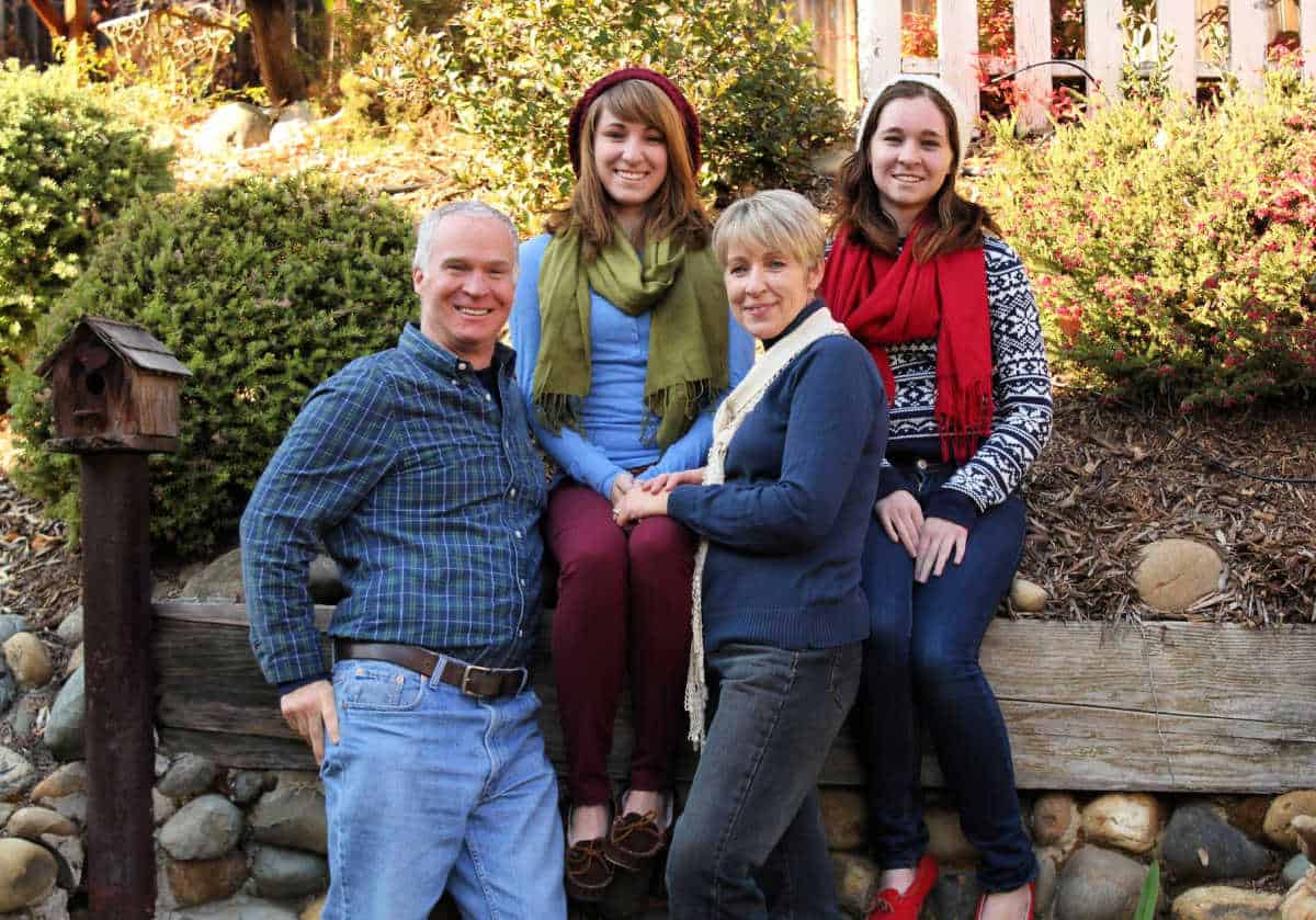 A man, two women, and a young woman pose together outdoors by a wooden beam and shrubbery, all smiling at the camera.