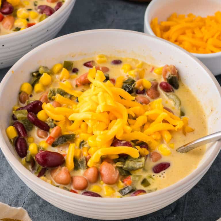 A bowl of Mexican corn and bean soup with kidney beans, pinto beans, corn, vegetables, and topped with shredded cheddar cheese; a spoon rests inside the bowl.