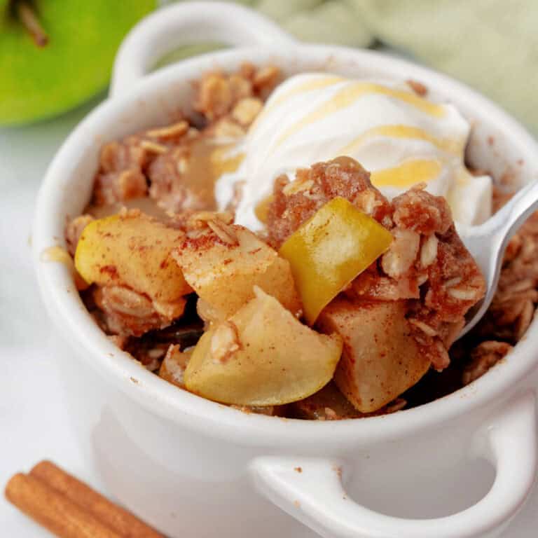 A white bowl filled with a single serve apple crisp topped with a scoop of vanilla ice cream and a drizzle of sauce, with a spoon inside.