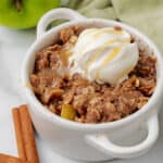 An apple crisp in a mug topped with whipped cream and caramel drizzle, with two cinnamon sticks and a green apple in the background.